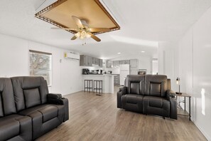 Un salon moderne avec un canapé et un fauteuil inclinable, un coin cuisine doté de placards et d’un plan de travail, et un ventilateur de plafond.