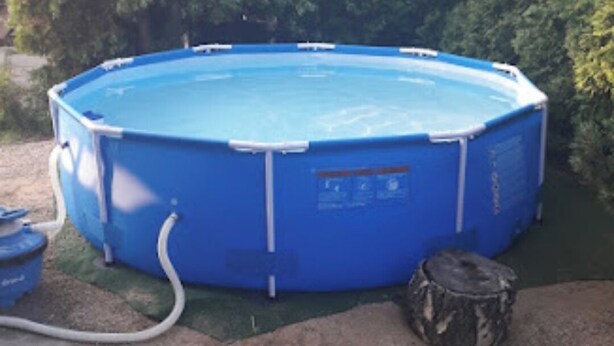 A blue above-ground swimming pool with a hose connected to it, surrounded by a green lawn and trees in the background.