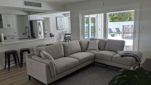A modern living room with a sectional sofa, a kitchen area with a refrigerator, and a view of a backyard with a pool. - Newly Renovated, Modern, Pristine w/ Heated Pool - 10 min to Siesta Beach (Sarasota)