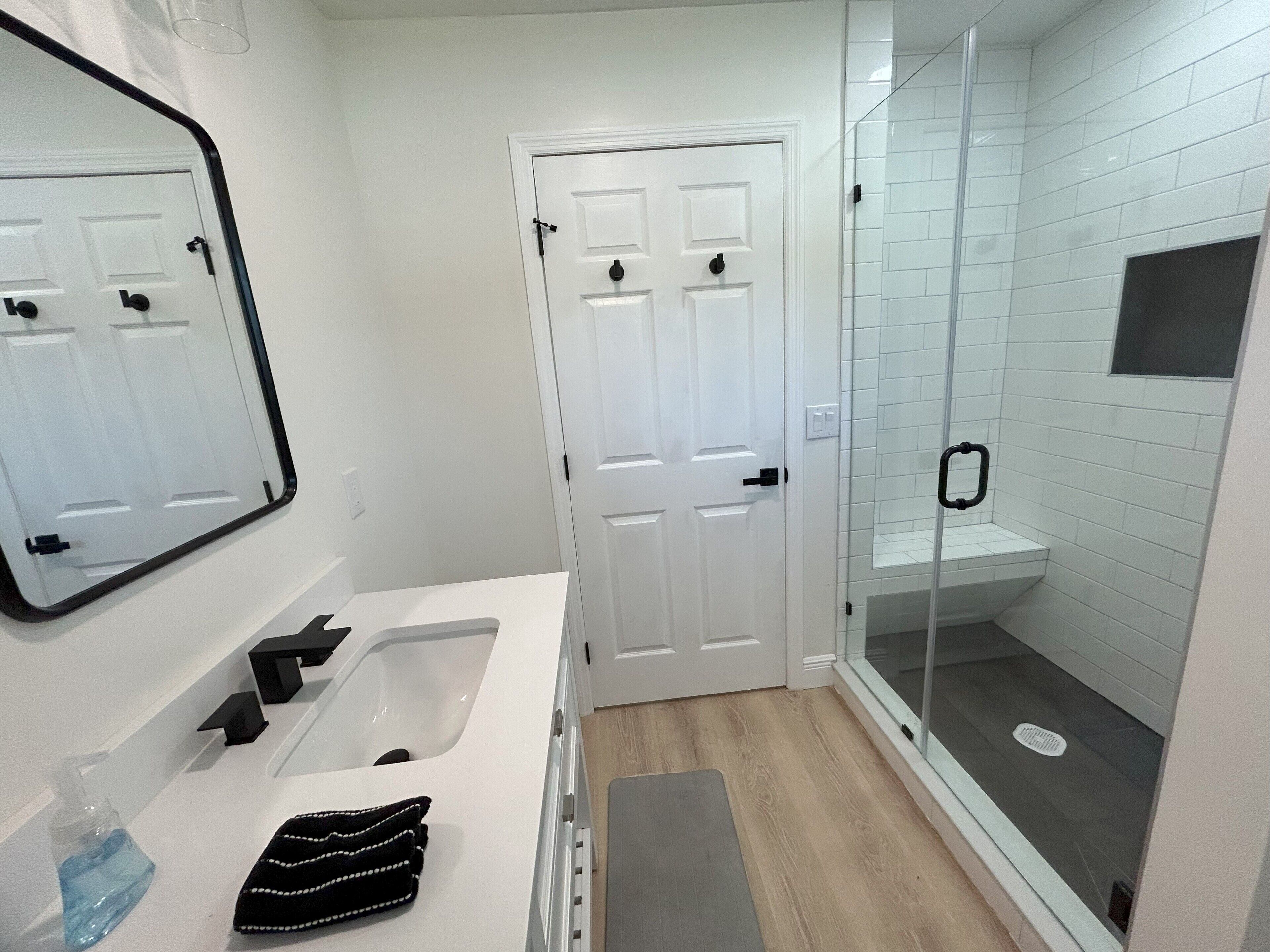 Un baño con lavamanos blanco, espejo y puerta, ducha con puertas de vidrio y una ventana pequeña.
