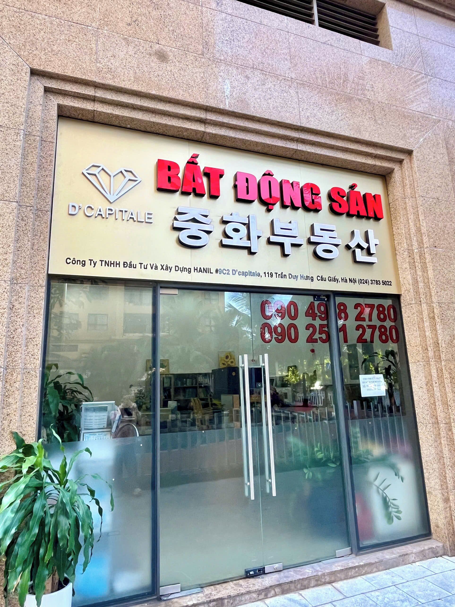 Ein Schaufenster mit einer großen Glasscheibe, einem Schild mit der Aufschrift 'BÁT ĐỘNG SÁN' und Kontaktnummern sowie einem Blumentopf davor.