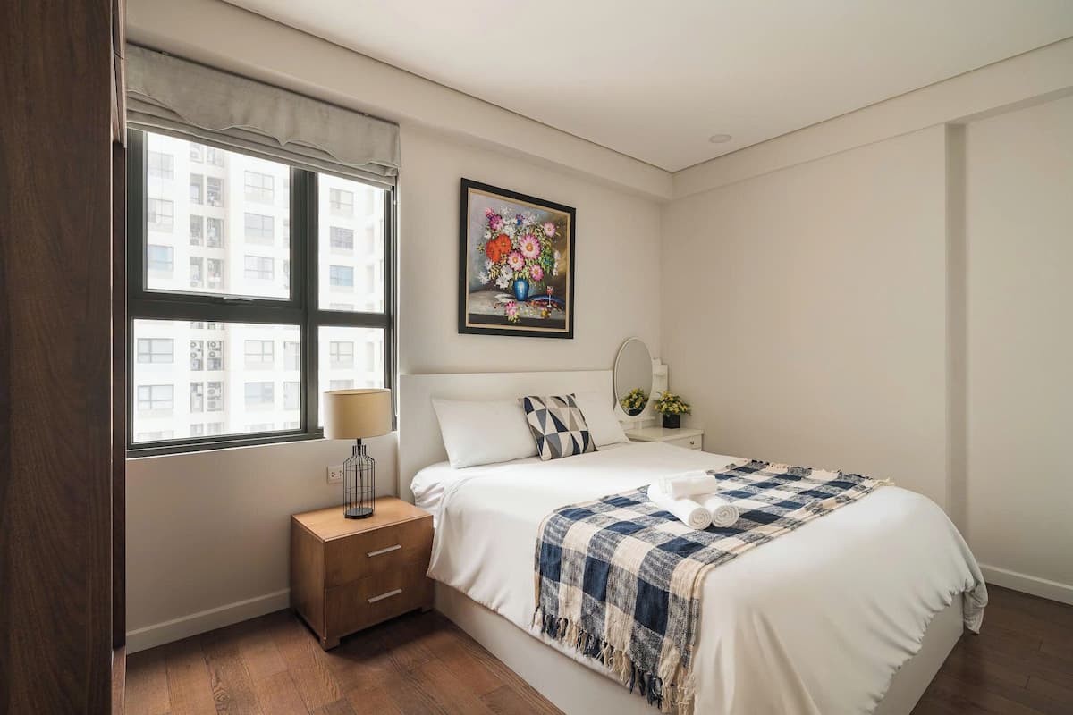 A bedroom with a bed, bedside table, lamp, and a window with a view of buildings.