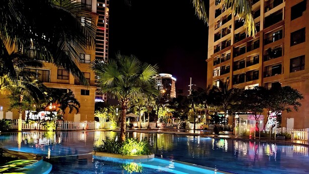 A nighttime pool area with a clear water surface, surrounded by palm trees and buildings. - Hopo Apt - Vinhomes D'capitale Luxury (Hanoi)
