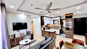 A modern living room with a TV, seating, and a kitchen area with a dining table. - Hopo Apt - Vinhomes D'capitale Luxury (Hanoi)