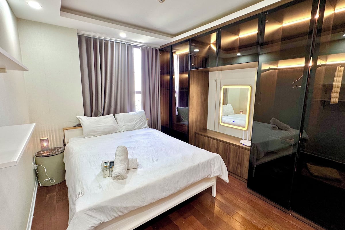 A modern hotel room with a large bed, a bedside table with a lamp, a mirror, and a wooden floor.