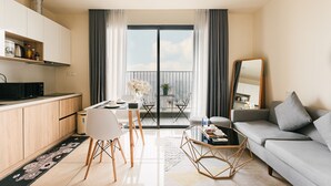 A modern living room with a sofa, a dining table with chairs, a kitchen area with cabinets, and a balcony with a view. - Hopo Apt - Vinhomes D'capitale Luxury (Hanoi)
