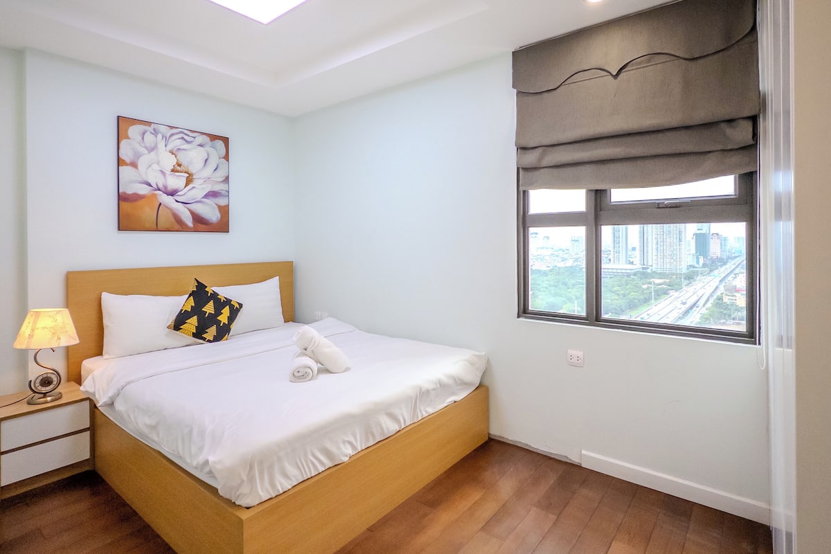 A bedroom with a bed, a nightstand, a lamp, a painting, and a window with a view of a cityscape.