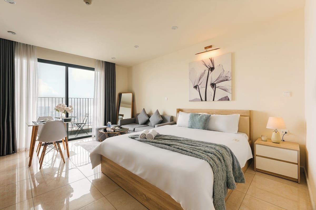 A modern hotel room with a large bed, a desk with a chair, a balcony with a view, and a painting on the wall.