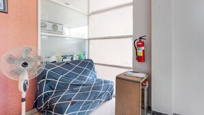 Une pièce comprenant un lit, un ventilateur sur pied, une petite table en bois, un extincteur et une fenêtre avec des stores.