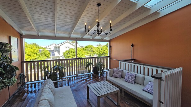 Una veranda riparata con un pavimento in legno, un divano bianco con cuscini decorati, un tavolo in legno e un soffitto con lucernari.