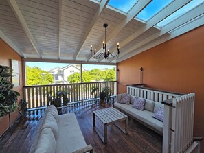 Eine überdachte Terrasse mit Holzdeck, einem weißen Sofa mit gemusterten Kissen, einem Holztisch und einer Decke mit Oberlichtern.
