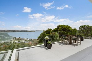 A balcony with a view of a lake, outdoor furniture, and potted plants. - Clifftop Glasshouse with Sweeping Views (Whangaparāoa)