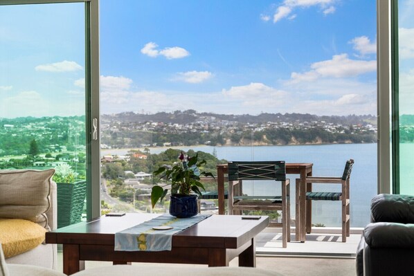 A balcony with a view of a lake and town, furnished with a table, chairs, and a potted plant. - Clifftop Glasshouse with Sweeping Views (Whangaparāoa)