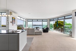 A spacious living area with a large window overlooking a body of water, a modern kitchen with a central island, and a cozy seating area with a sofa and armchair. - Clifftop Glasshouse with Sweeping Views (Whangaparāoa)