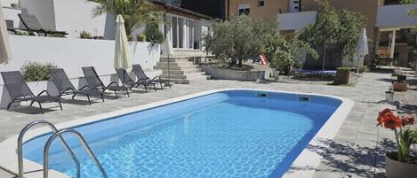 A rectangular swimming pool with a tiled deck, surrounded by lounge chairs and a house with a tiled roof.