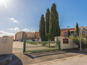 Ein befestigter Eingang zu einem Gebäude mit Parkplatz, hoch aufragenden Zypressen und blauem Himmel.