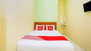 Habitación de hotel estrecha con una cama, dos almohadas, una mesita de noche y un televisor de pared.