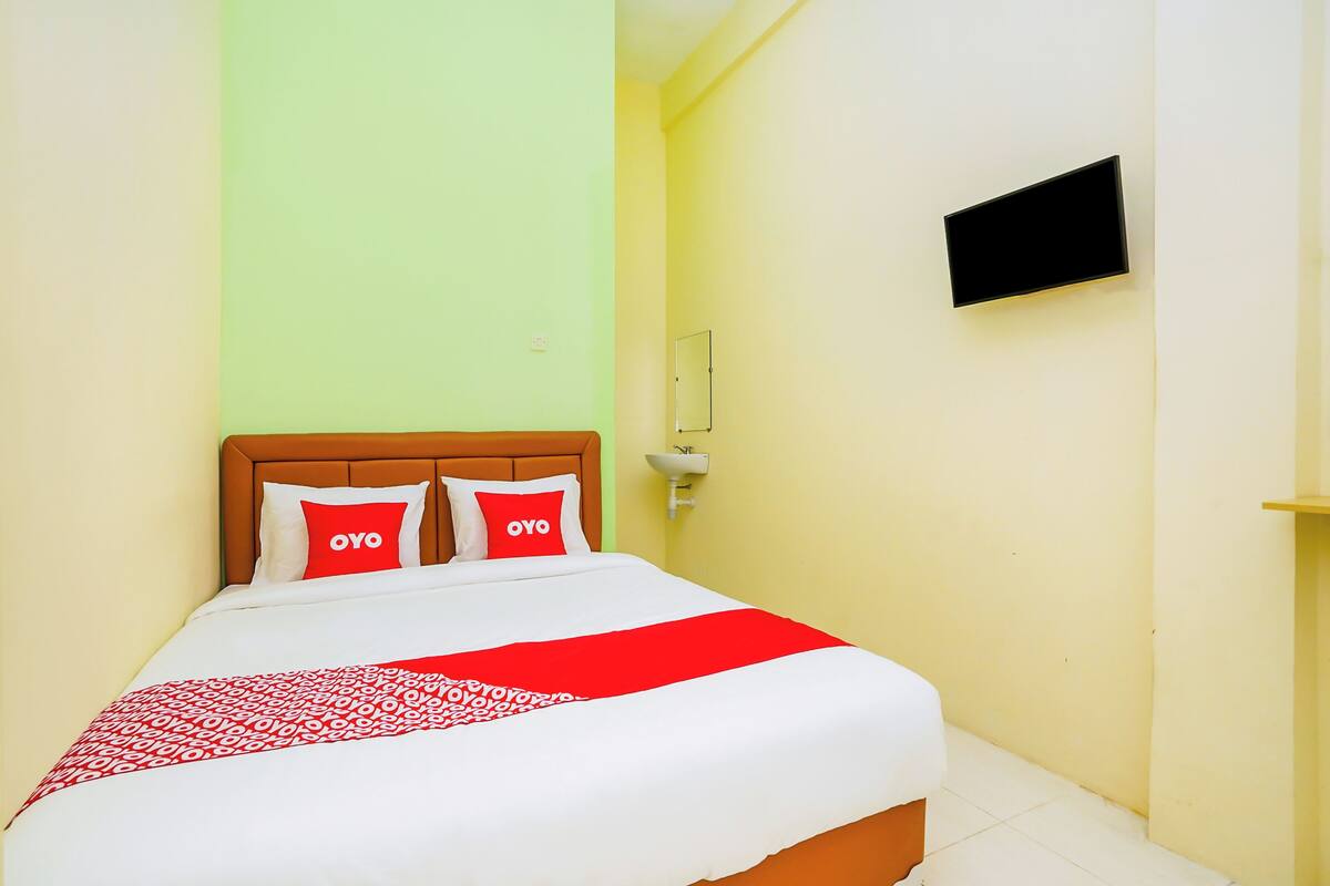 Habitación de hotel con cama, dos almohadas con la palabra 'OYO', una colcha estampada en rojo y blanco, un televisor montado en la pared y un pequeño estante.