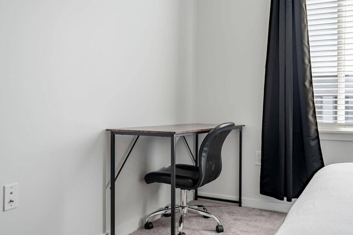 Une petite pièce, bien rangée, avec un bureau et une chaise, une fenêtre avec des stores et un lit.