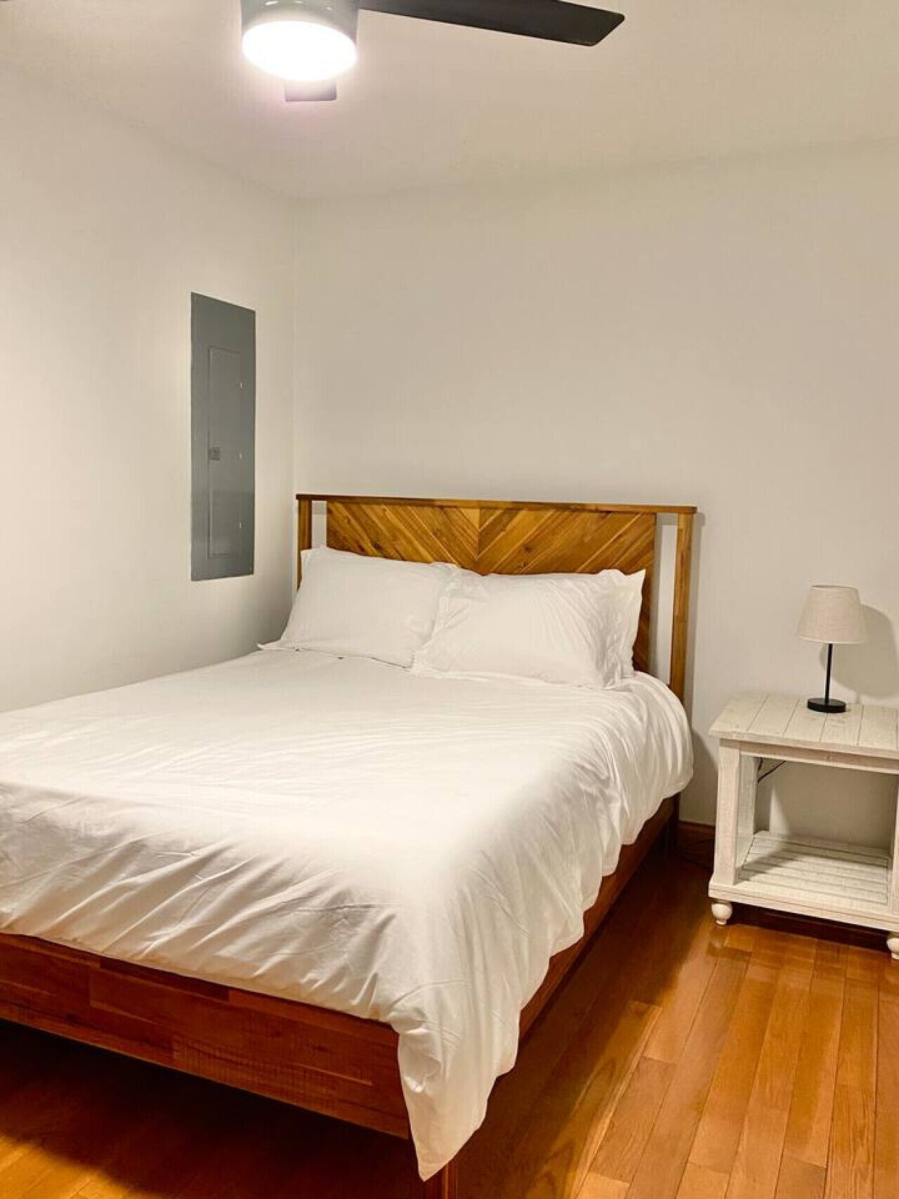 A bedroom with a wooden bed frame, white bedding, a nightstand with a lamp, and a wall-mounted light fixture.