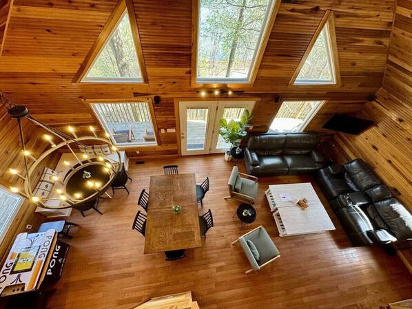 A cozy cabin interior with a wooden floor, a central dining table surrounded by chairs, a sofa, and a large window providing natural light.