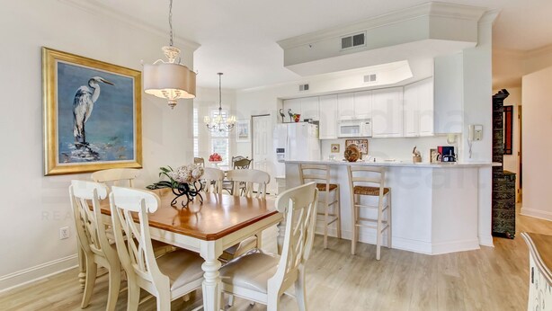 A modern kitchen with white cabinetry, a dining area with a wooden table and white chairs, and a painting of a bird on the wall.