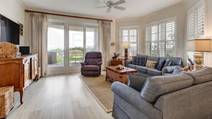 A spacious living room with a sofa, armchair, and a wooden coffee table. There is a ceiling fan, a TV mounted on the wall, and large windows with blinds.