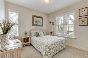 A bedroom with a bed, a bedside table, a lamp, a window with blinds, and wall decorations. - 75 Ocean Place (Fernandina Beach)