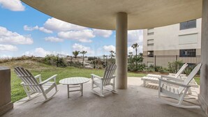 A covered outdoor seating area with white Adirondack chairs and a round table, overlooking a grassy area and a building with multiple windows.