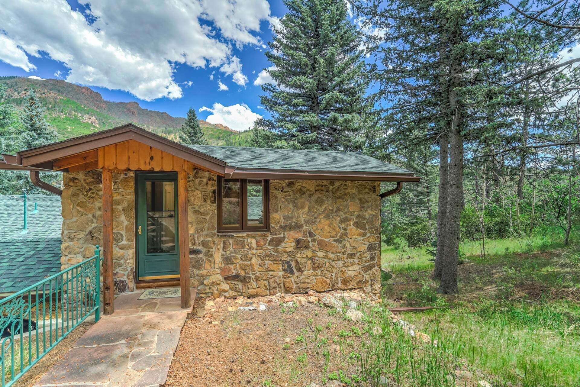 Une cabane en pierre avec une porte verte, un petit porche et une vue sur une montagne boisée.