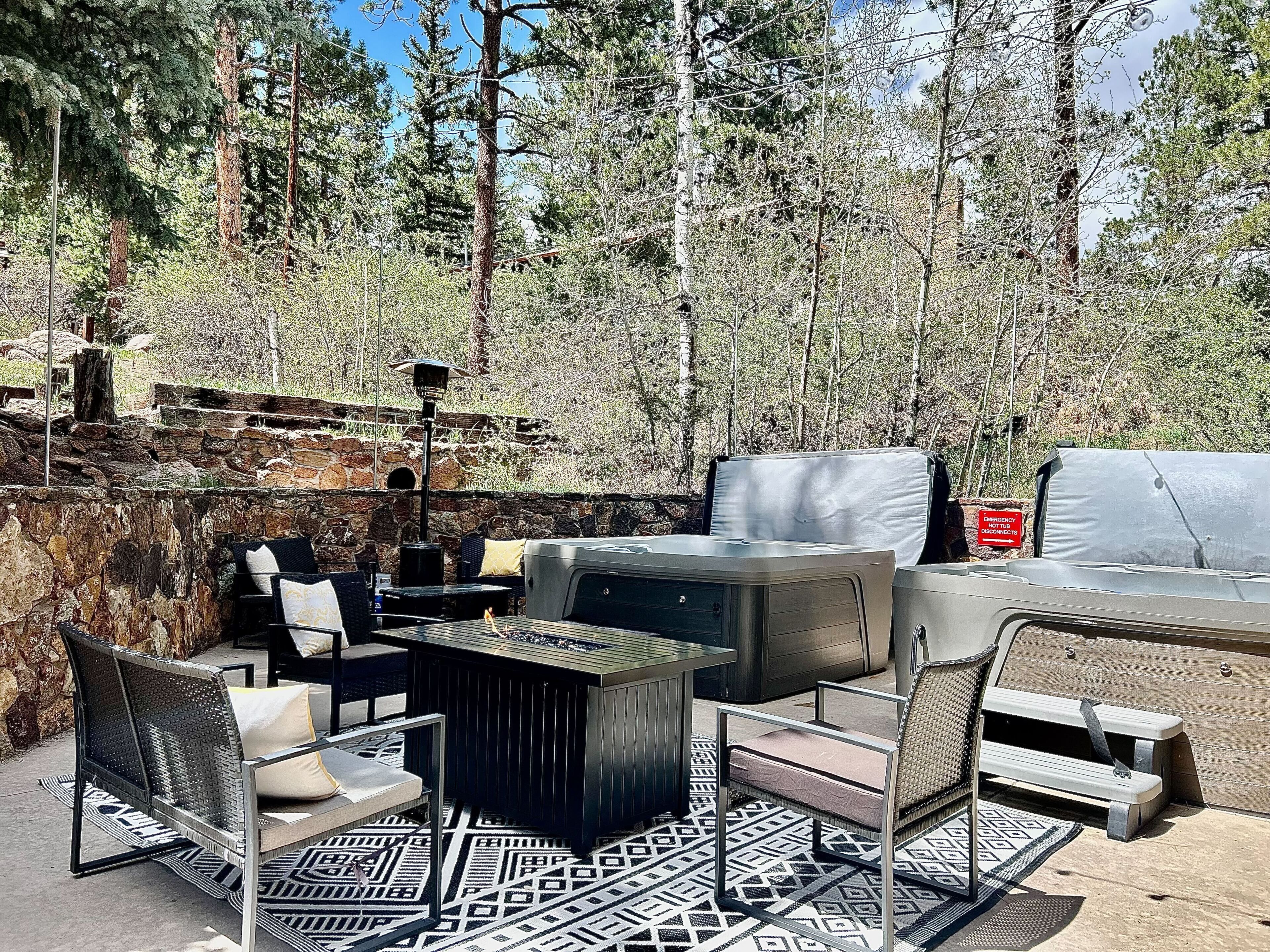 An outdoor seating area with a grill, chairs, and a table, surrounded by trees and a stone wall.
