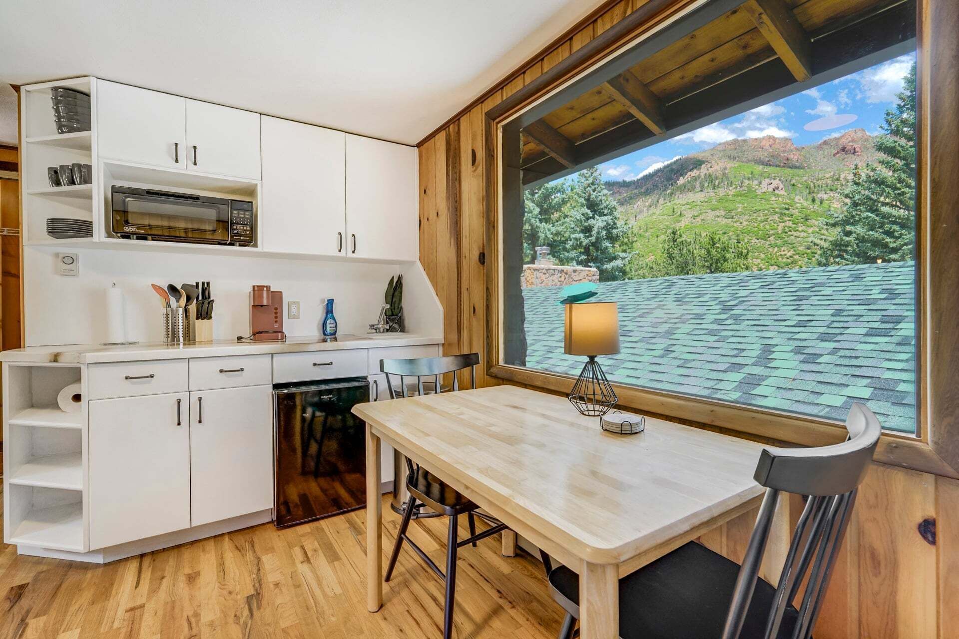 A compact kitchen with white cabinets, a microwave, and a coffee maker. There's a dining table with chairs, and a large window offering a view of a mountainous landscape.