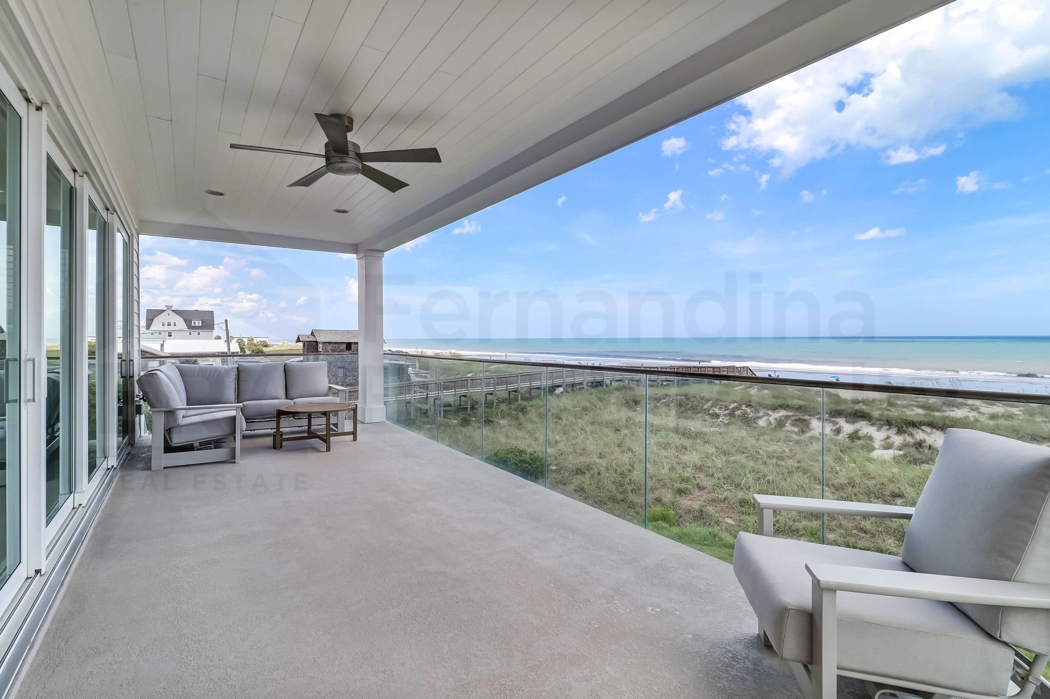 A covered balcony with a ceiling fan, a sofa, a chair, and a view of the beach and dunes.