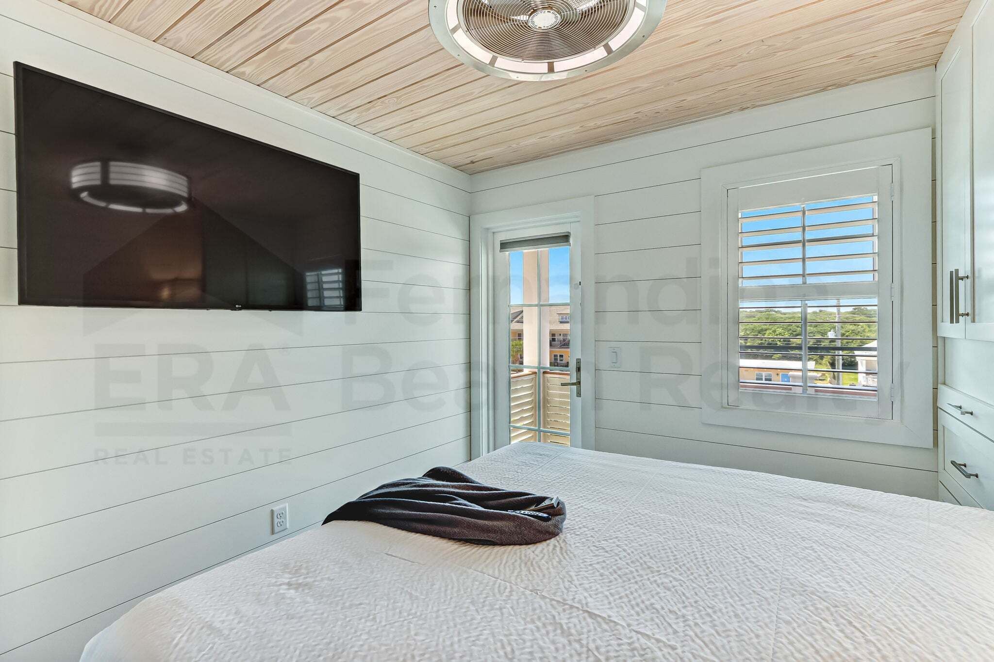 A bedroom with a bed, a TV mounted on the wall, a ceiling fan, and a window with blinds.