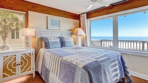 Een slaapkamer met een groot bed, houten balken in het plafond, uitzicht op de oceaan en een witte nachtkastje met een lamp.