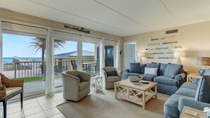 A spacious living room with a large sliding glass door leading to a balcony, a blue sofa set, a wicker chair, and a small coffee table. - 108 Amelia by the Sea (Fernandina Beach)