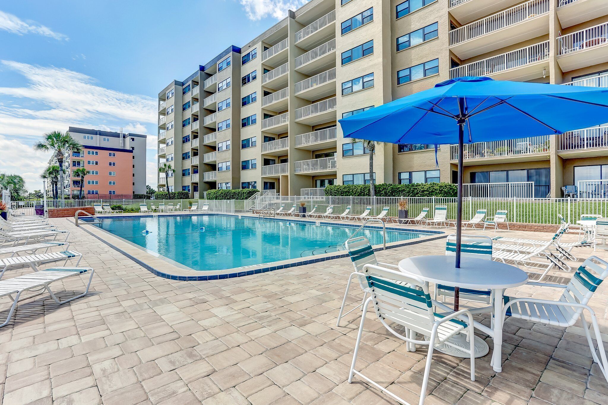 A pool area with lounge chairs and a table, surrounded by a fence and a multi-story building.