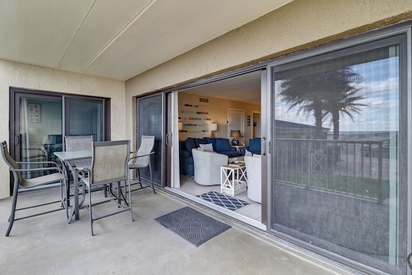 A patio with a table and chairs, a sliding glass door leading to an interior with a sofa and a lamp.
