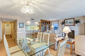 Een ruime woonkamer met een glazen eettafel, witte stoelen, een open haard en een keuken op de achtergrond.