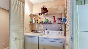 A laundry room with two stacked washers and dryers, shelves above holding cleaning supplies, and a white refrigerator to the right. - 301 Atlantis (Fernandina Beach)
