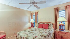 A bedroom with a bed, nightstands, a lamp, a ceiling fan, and a window with curtains. - 301 Atlantis (Fernandina Beach)