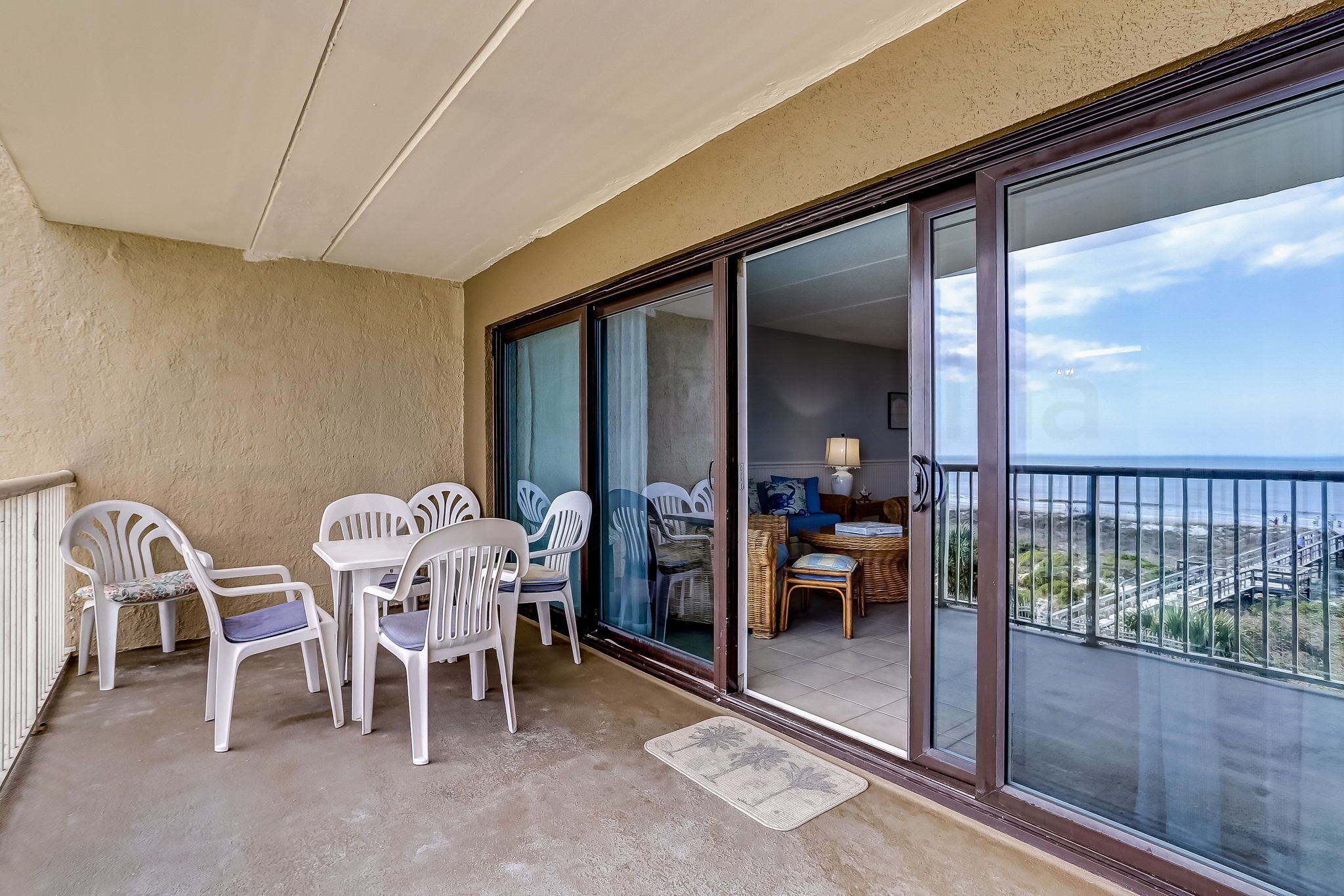 A balcony with white plastic chairs and a table, a sliding glass door leading to an interior space with a wicker table and chairs, and a view of the coastline.
