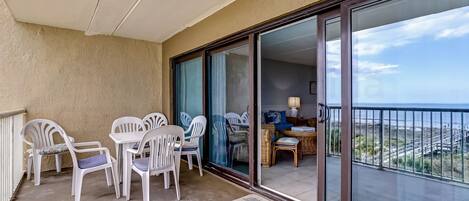 A balcony with white plastic chairs and a table, a sliding glass door leading to an interior space with a wicker table and chairs, and a view of the coastline.