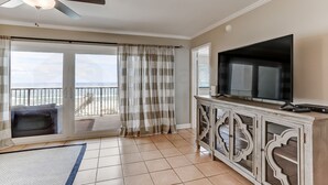 A room with a large TV, a ceiling fan, a balcony with curtains, and a view of the ocean. - 6th Floor, Updated and Beautiful! Oceanfront Condo With Balcony, Fishing Pier (Fernandina Beach)