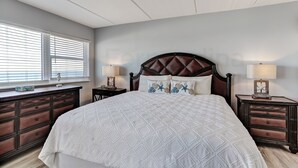 A bedroom with a large bed, dark wooden headboard, two bedside tables with lamps, a dresser, and a window with blinds.