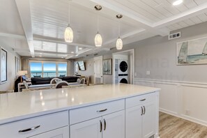 Une cuisine moderne avec des meubles blancs, un grand îlot central, des luminaires suspendus et une vue sur l’océan.