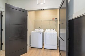 Une buanderie avec deux machines à laver blanches, une étagère en métal contenant des produits de lessive et une porte sombre.