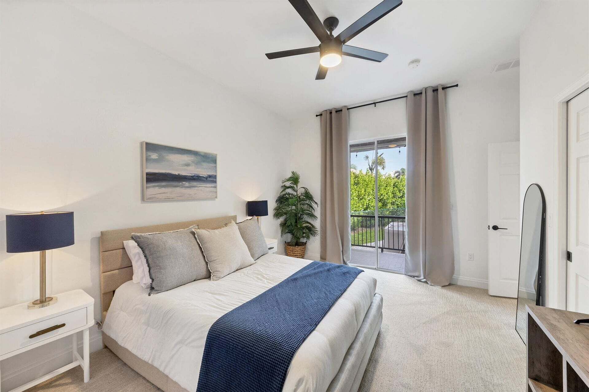 Une chambre à coucher comprenant un lit, des tables de chevet, un ventilateur de plafond, une fenêtre avec des rideaux et une vue sur la verdure à l’extérieur.