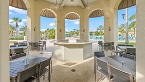 Un espace de restauration extérieur abrit é, avec des tables et des chaises, un îlot central et une vue sur une piscine et des palmiers.