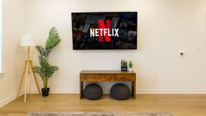 A modern living room with a large Netflix logo on the wall, a wooden bench with storage, a potted plant, and a floor lamp. - Relaxing Getaway, Worker-friendly, A+ Location (Sherman)